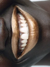 Extreme close-up macro portrait of dark-skinned lips painted with metallic bronze lipstick, slightly parted to reveal white teeth.