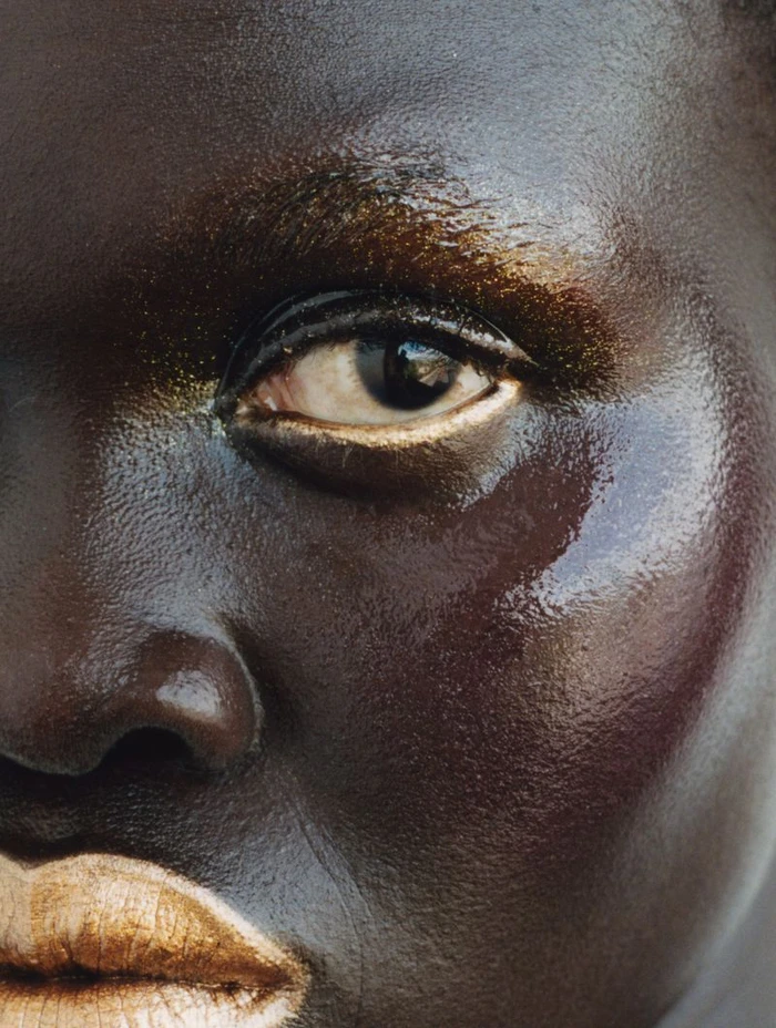 Extreme close-up macro portrait of a person with dark skin showing gold glitter makeup around the eye and lip