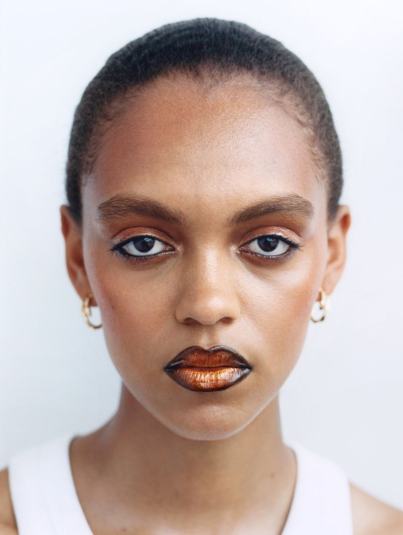 Close-up portrait of a young adult female with bronze eyeshadow and metallic lips in a bright studio against a white background.