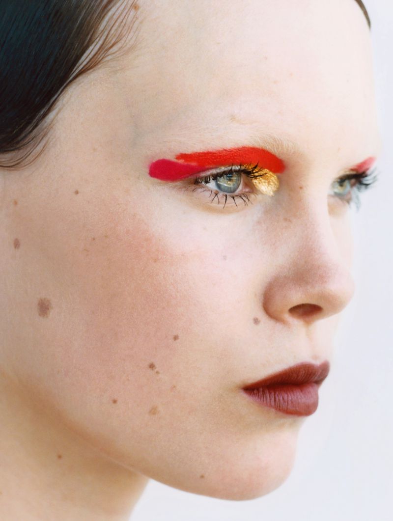 Close-up portrait of a fair-skinned young adult woman in profile with creative makeup: red eyeliner and dark red lipstick against a white background.