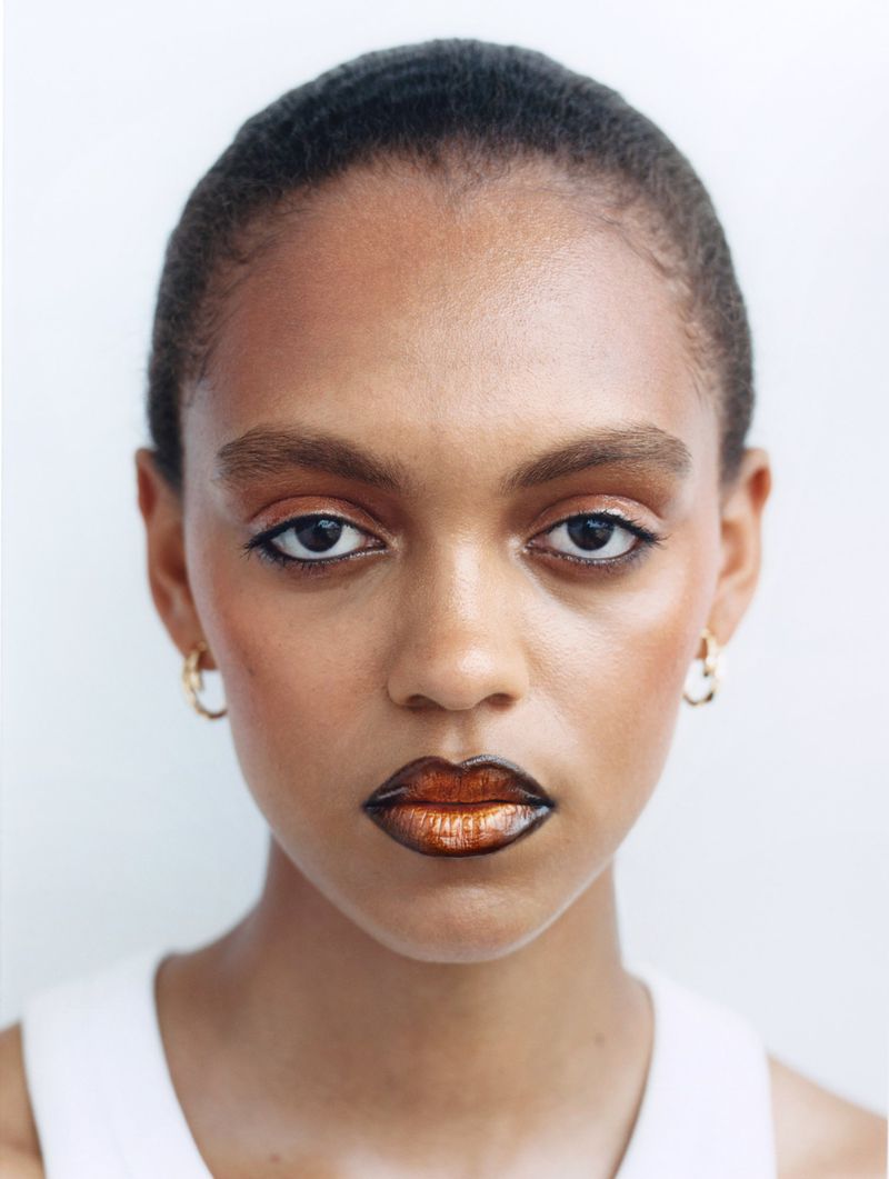 Close-up studio portrait of an adult Black woman with gold eye and lip makeup, hoop earrings, and a neutral facial expression.