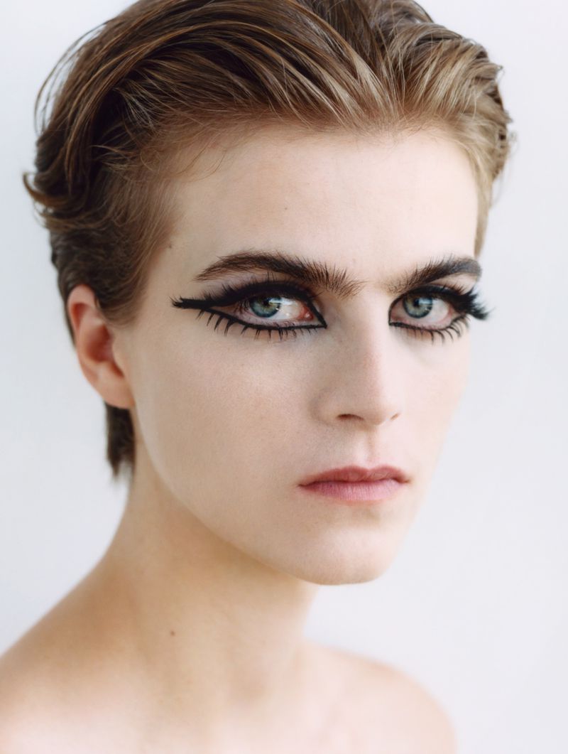Close-up portrait of a fair-skinned young adult with short brown hair and bold black winged eyeliner against a plain white background.