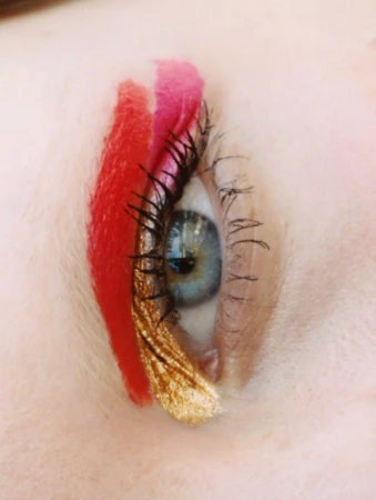 Close-up of a female eye with creative makeup, including red, pink, and gold accents, with visible eyelashes and fair skin tone.