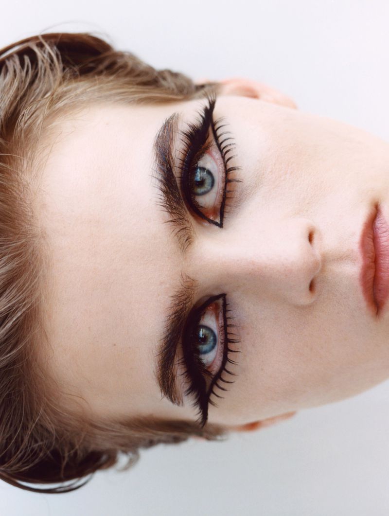 Close-up studio portrait of a fair-skinned young woman with dramatic black eyeliner and false eyelashes against a white background.