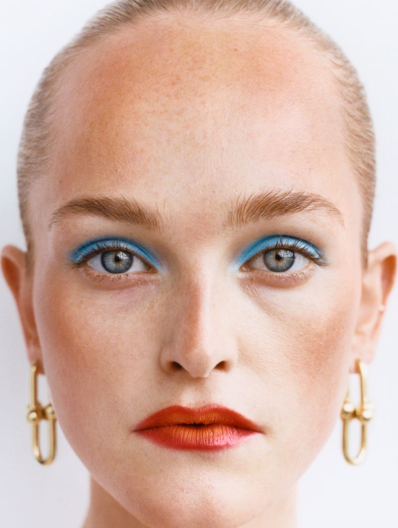 Close-up portrait of a fair-skinned woman with bright blue eyeshadow, vibrant red lipstick, and gold jewelry against a white backdrop.