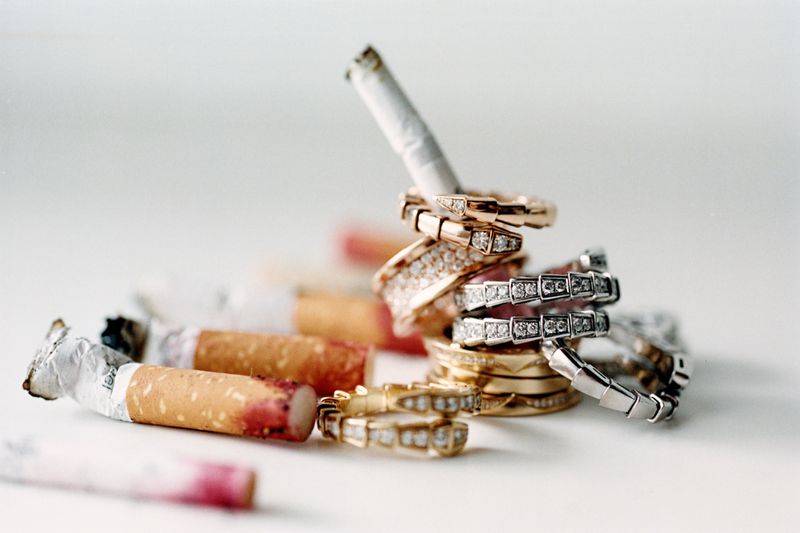 Conceptual still life composition featuring several gold and silver rings stacked around unlit and used cigarettes on a white surface.
