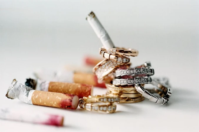 Conceptual still life composition featuring several gold and silver rings stacked around unlit and used cigarettes on a white surface.