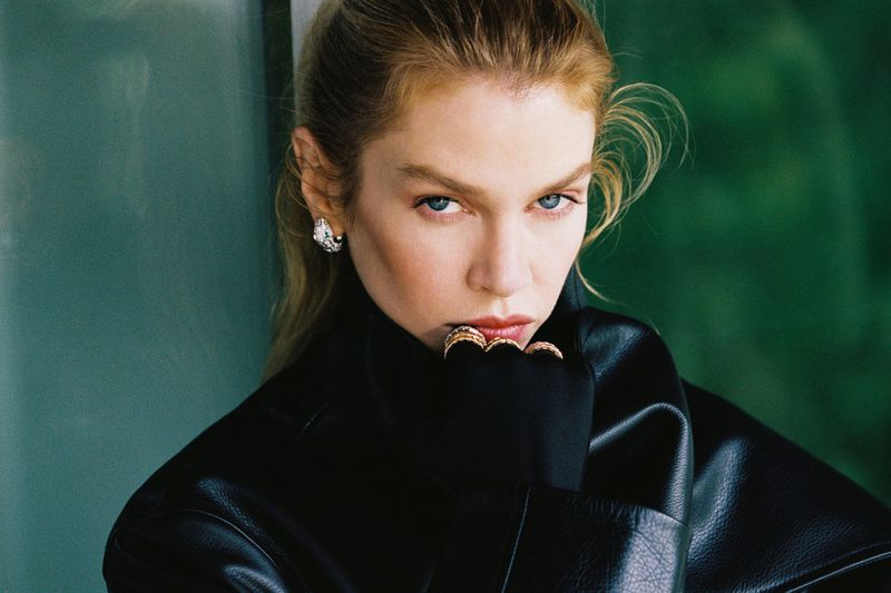 Model Stella Maxwell wearing a black leather jacket and black turtleneck with rings near her lips in a close-up portrait.