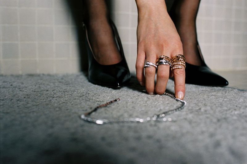 Close-up shot of a woman's hand with rings, touching a silver necklace on a carpeted floor next to a pair of black high heels.