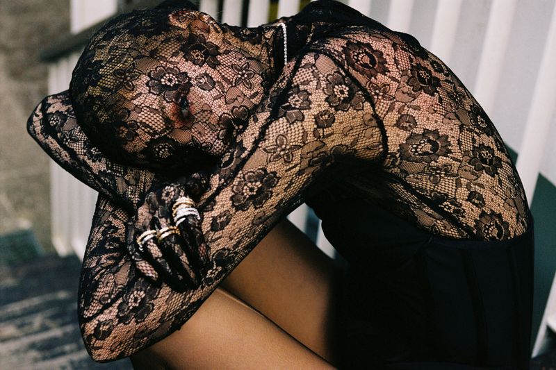 A person in a black lace bodysuit sits on steps, embracing their knees with arms covered in sheer black floral lace fabric.
