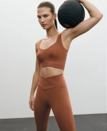 Model holding a black exercise ball on her shoulder, wearing rust activewear against a white studio background.