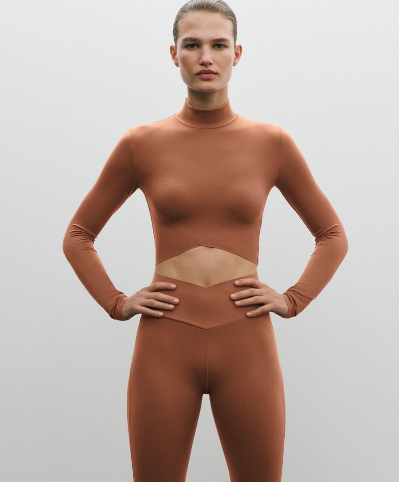 Model wearing a brown activewear set with a long-sleeve crop top and leggings against a seamless grey background