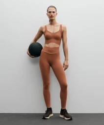 Model in rust-brown sports bra and leggings holding a medicine ball against a neutral grey studio background