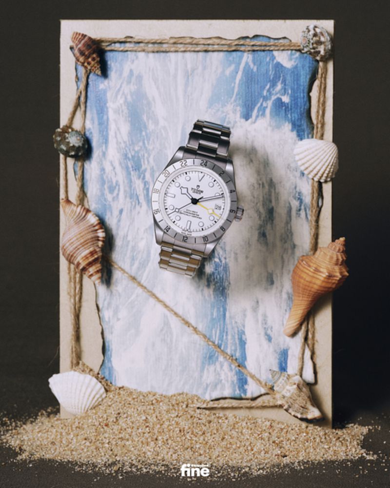 Luxury wristwatch displayed on a sandy surface with seashells and ocean wave art in the background in a staged product shot.