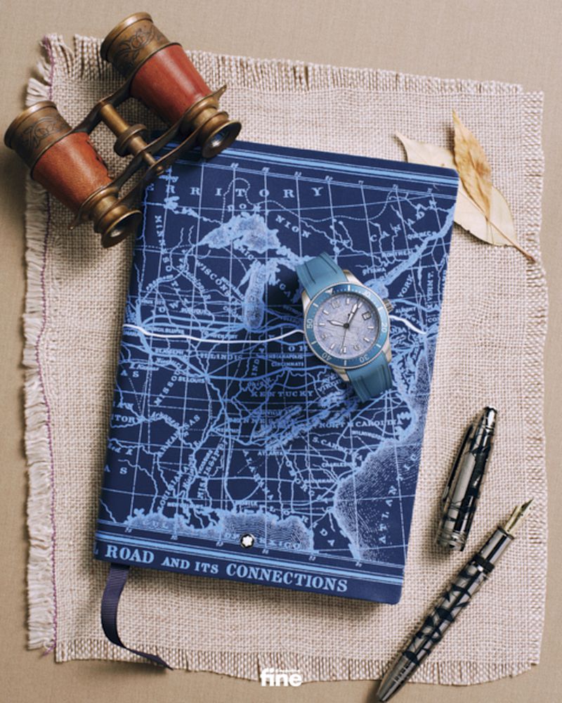 Elevated shot of a navy notebook with a world map, a wristwatch, a fountain pen, and binoculars on a textured neutral surface.