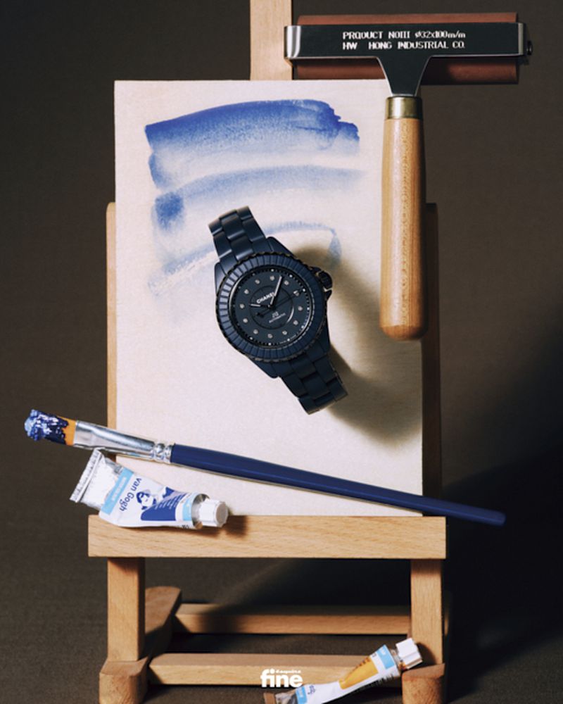 Still life of a black Chanel watch on an easel with blue watercolor paint, a paintbrush, and tubes of paint in a studio setting.