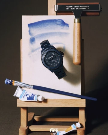 Still life of a black Chanel watch on an easel with blue watercolor paint, a paintbrush, and tubes of paint in a studio setting.