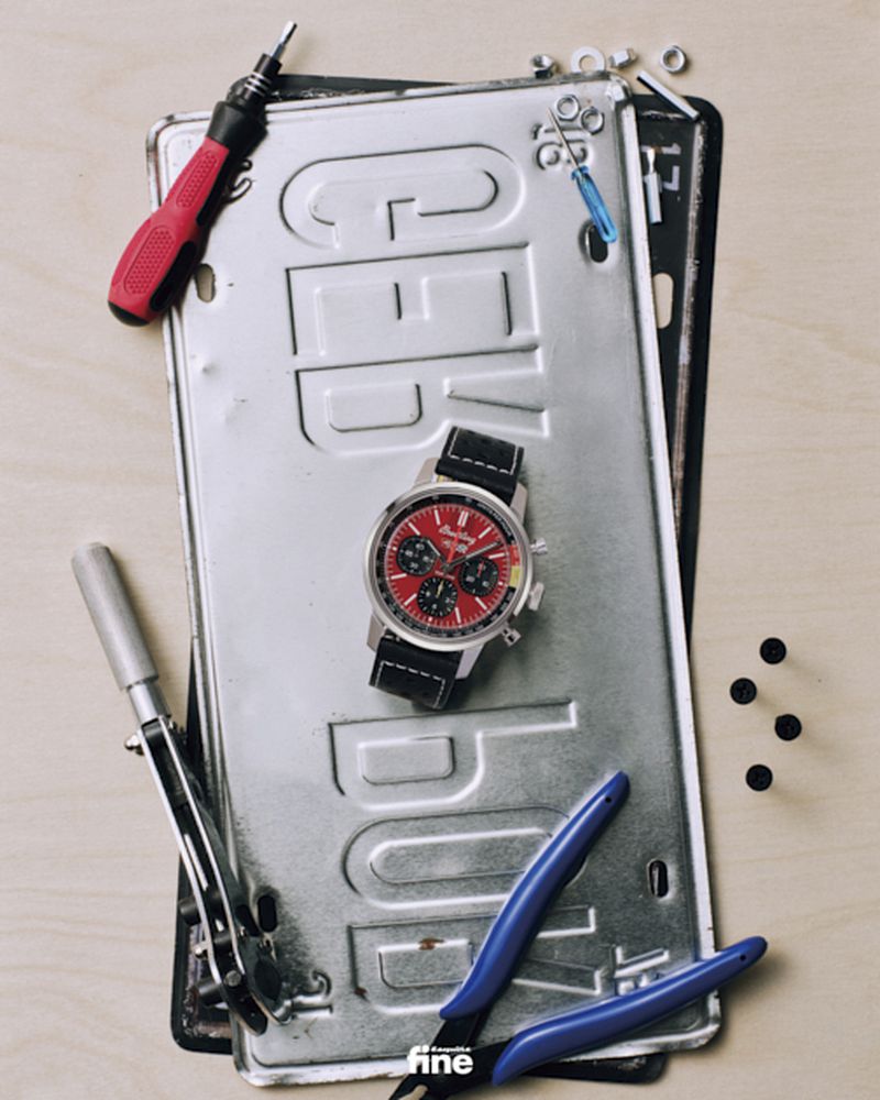 Analog watch with a red dial and black band is centered on top of a silver license plate surrounded by tools and hardware.