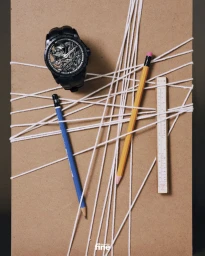 Black skeleton watch, two pencils, and a small ruler tangled in white string on a brown cardboard surface.