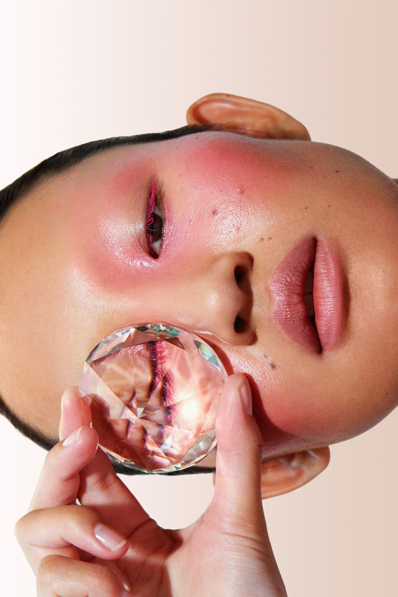 Artistic close-up portrait of a person with creative blush makeup, holding a clear crystal over the face, set against a neutral backdrop.