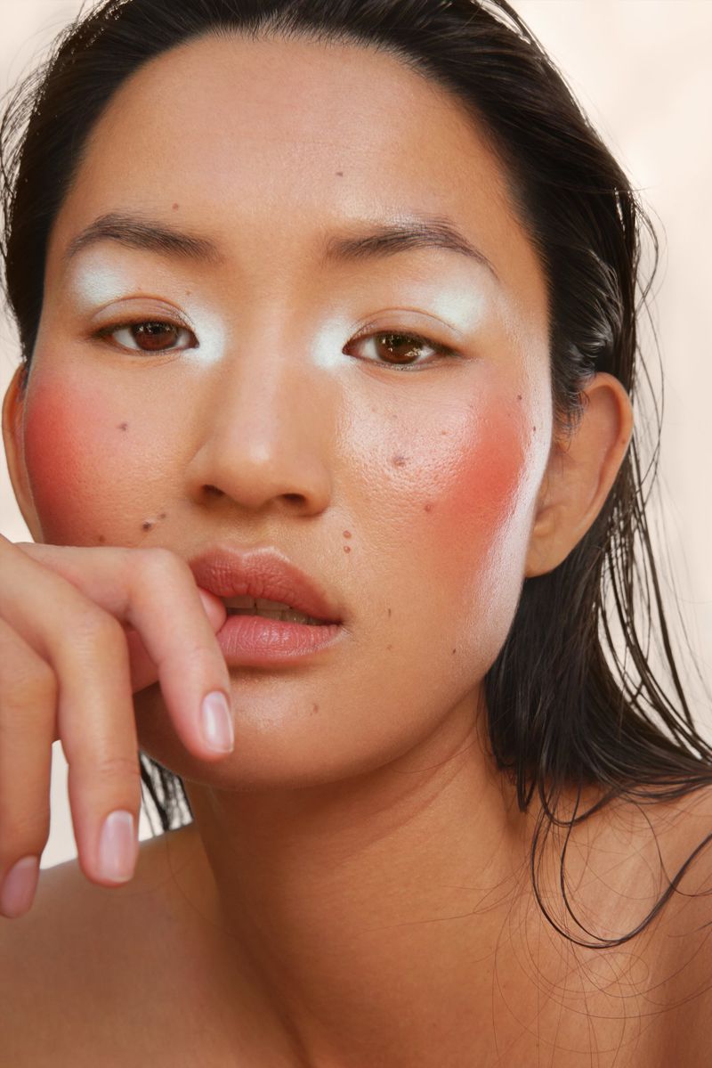 Close-up portrait of an Asian woman with wet hair, artistic makeup including light blue eyeshadow and rouge, set against a plain background.