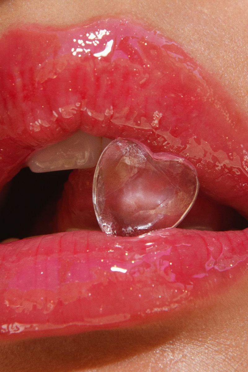 Close-up studio shot of pink lips with clear gloss holding a translucent heart-shaped ice cube. Focus on cosmetic beauty.