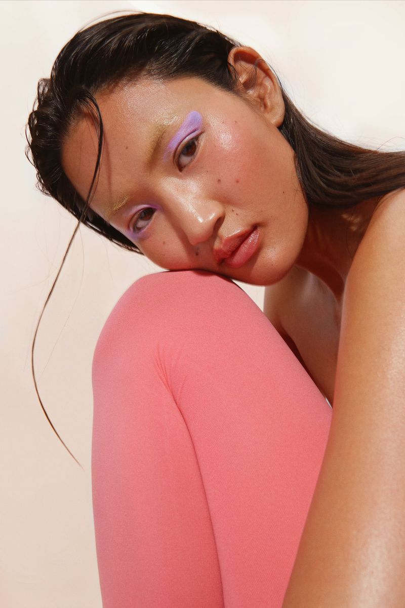 Close-up of an Asian woman with lavender and gold eye makeup posing in photo studio with wet hair and bright pink leggings.