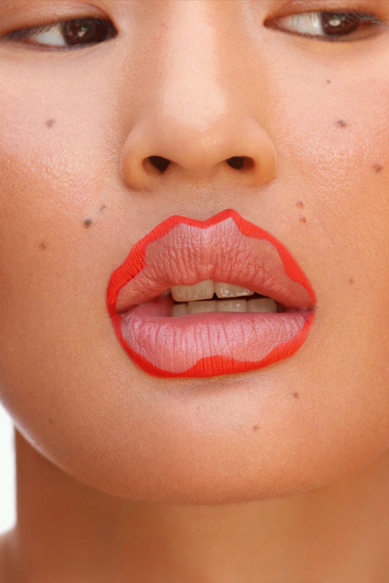 Close-up studio portrait of an Asian woman with freckles and red lipstick outlining her lips, neutral expression, and white background.