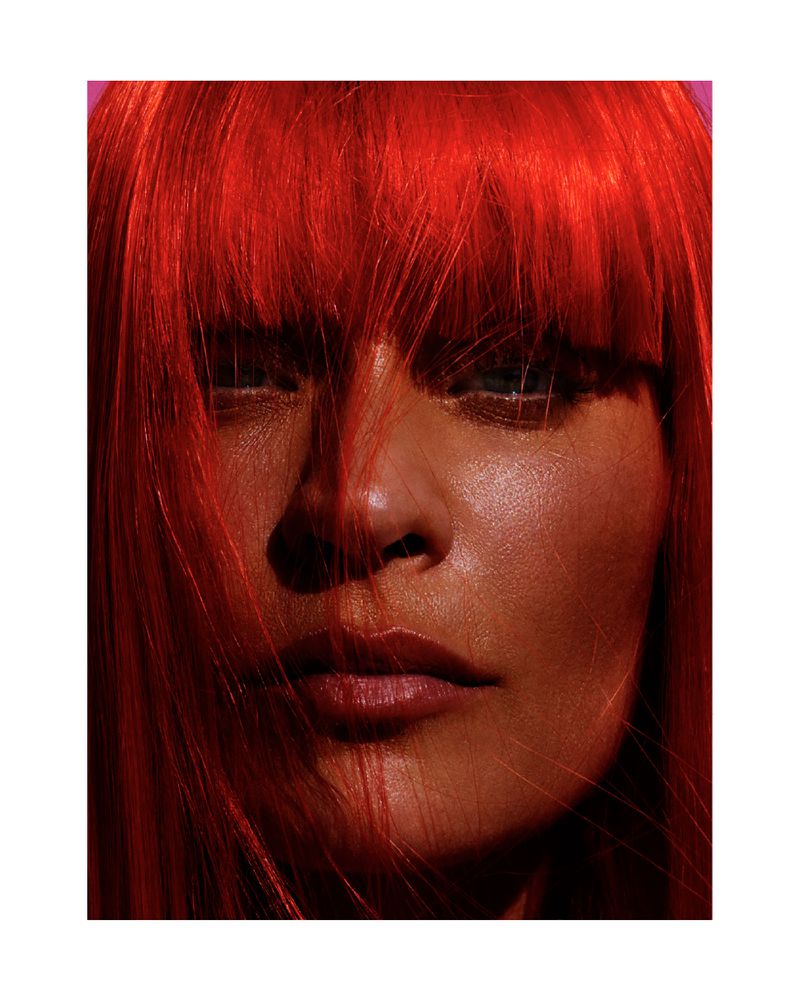 Close-up portrait of a woman with red hair and light skin, captured in warm, studio lighting against a magenta background.