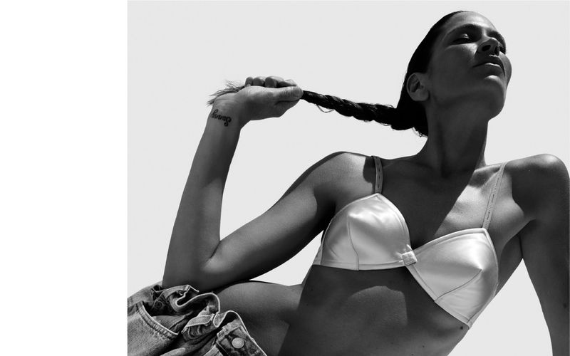 Monochrome portrait of a woman in a bra top and jeans, lying down, holding her hair, with natural light casting shadows on her skin.
