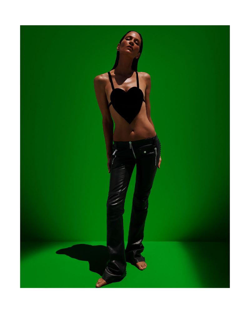 A slender woman in a heart-shaped top and leather pants poses against a vibrant green backdrop, illuminated by directed studio lighting.
