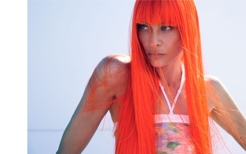 Woman with bright orange wig gazes off-camera in a fashion shot with a light blue background, floral-patterned top, and white straps.