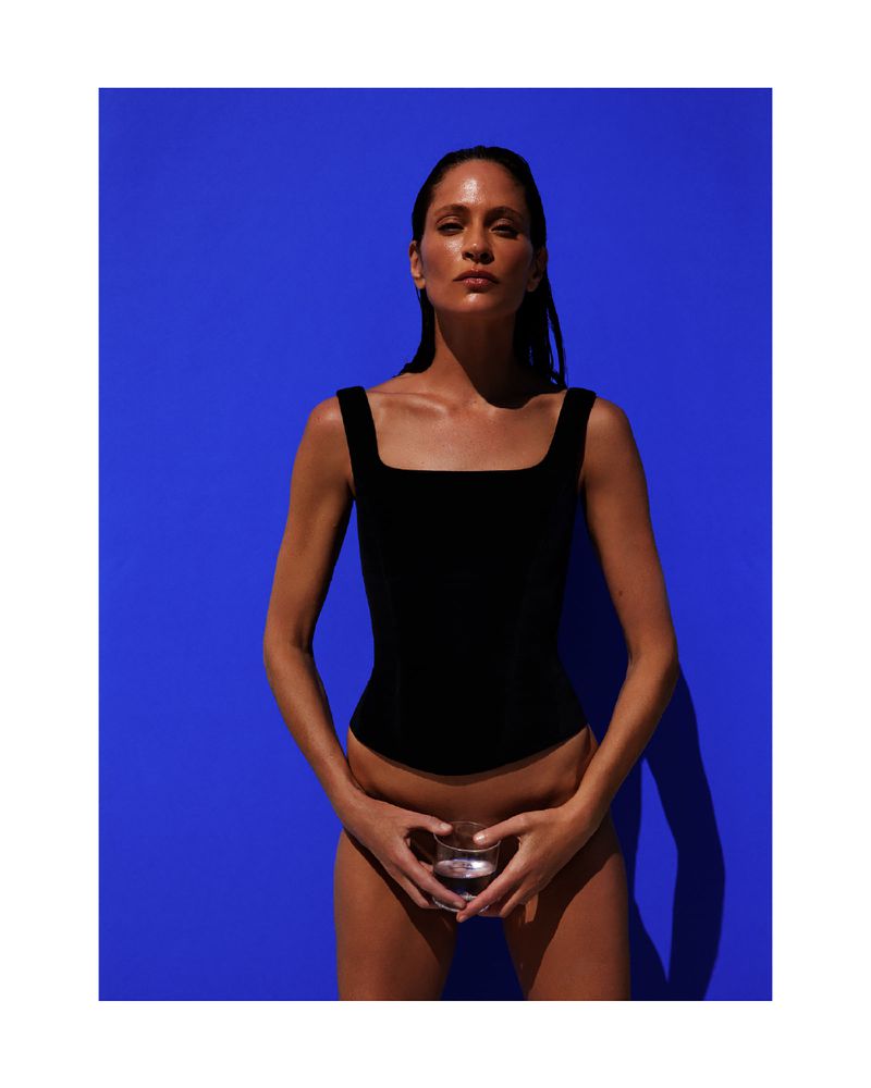A woman in a black bathing suit stands against a vibrant blue background, holding a glass of clear liquid with both hands in a studio shot.
