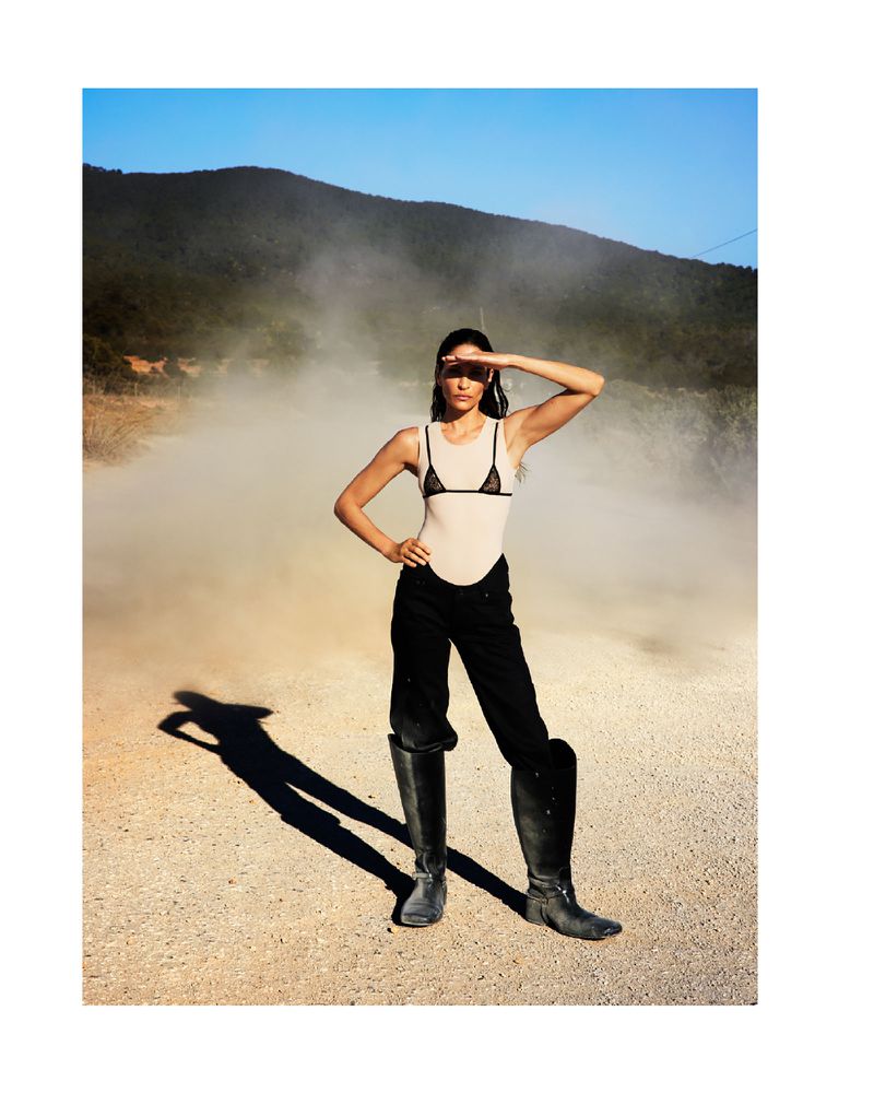 A woman in riding boots and bodysuit stands in a dusty, sunlit landscape with a mountain backdrop, shielding her eyes from the sun.