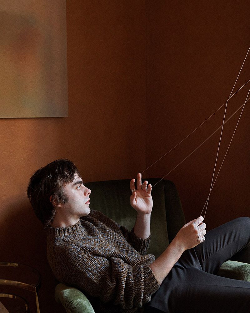 Man in brown sweater relaxes in green chair, holding strings. Interior shot featuring neutral tones and natural light.
