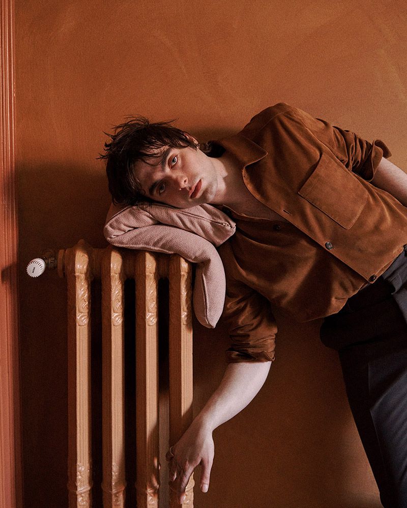 Portrait of a young adult male with dark hair, wearing a brown button-down shirt, resting his head on a pillow against a radiator indoors.