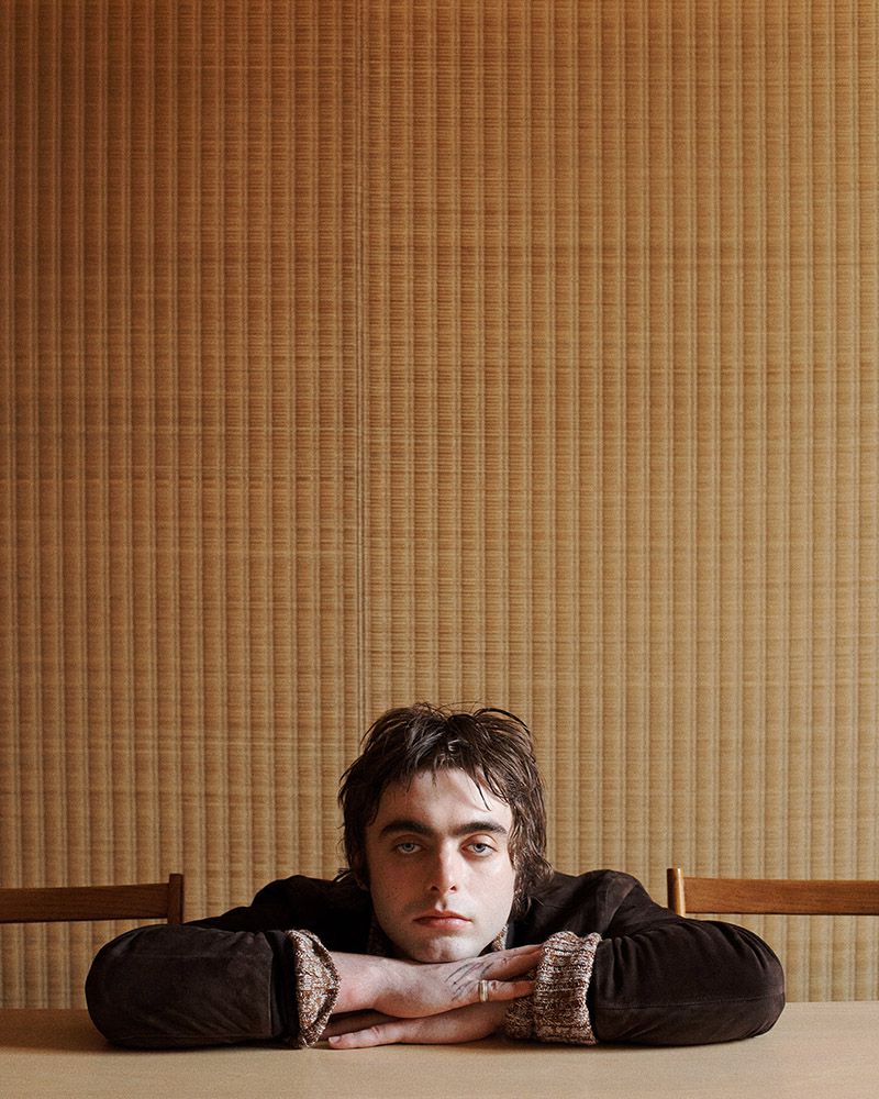 Portrait of a man with dark hair and green eyes leaning on a tan table against a light brown textured wall in an indoor setting.