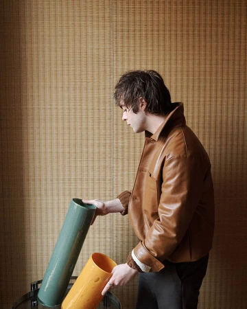 Man in brown jacket with two decorative vases, one green and one orange, in an indoor setting with textured beige wallpaper.