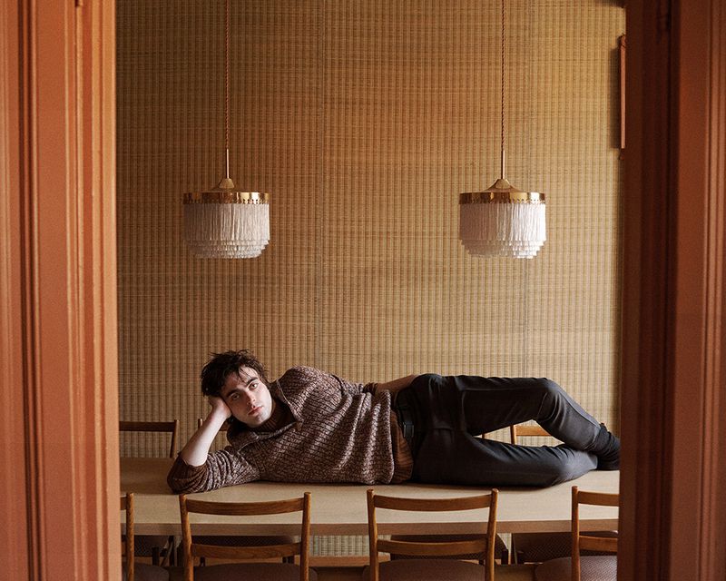 A relaxed adult male poses on a wooden table in a warm-toned indoor setting, with pendant lights and wood-slat wall visible.
