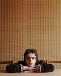 Portrait of model Lennon Gallagher leaning with crossed arms on a table against a textured brown wall background.