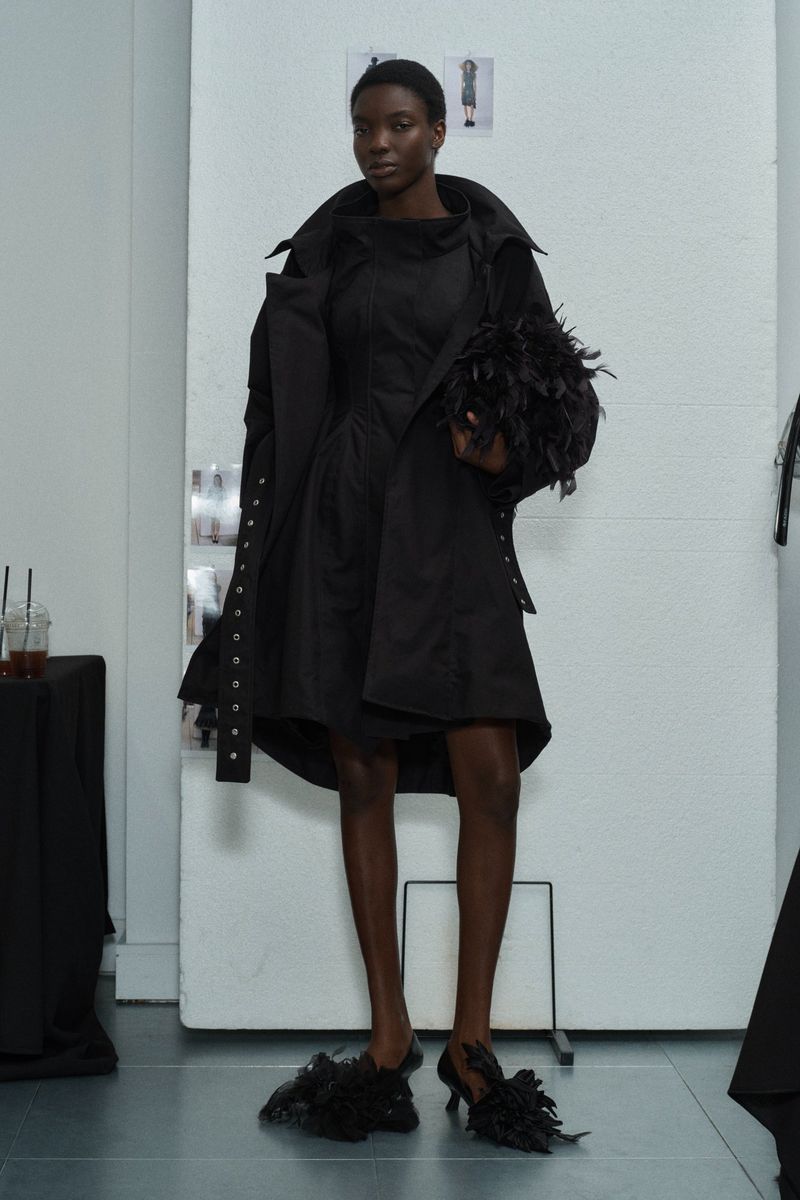 Runway fashion: A Black female model showcases a black dress and coat ensemble with feather-accented shoes and bag in a photo studio.
