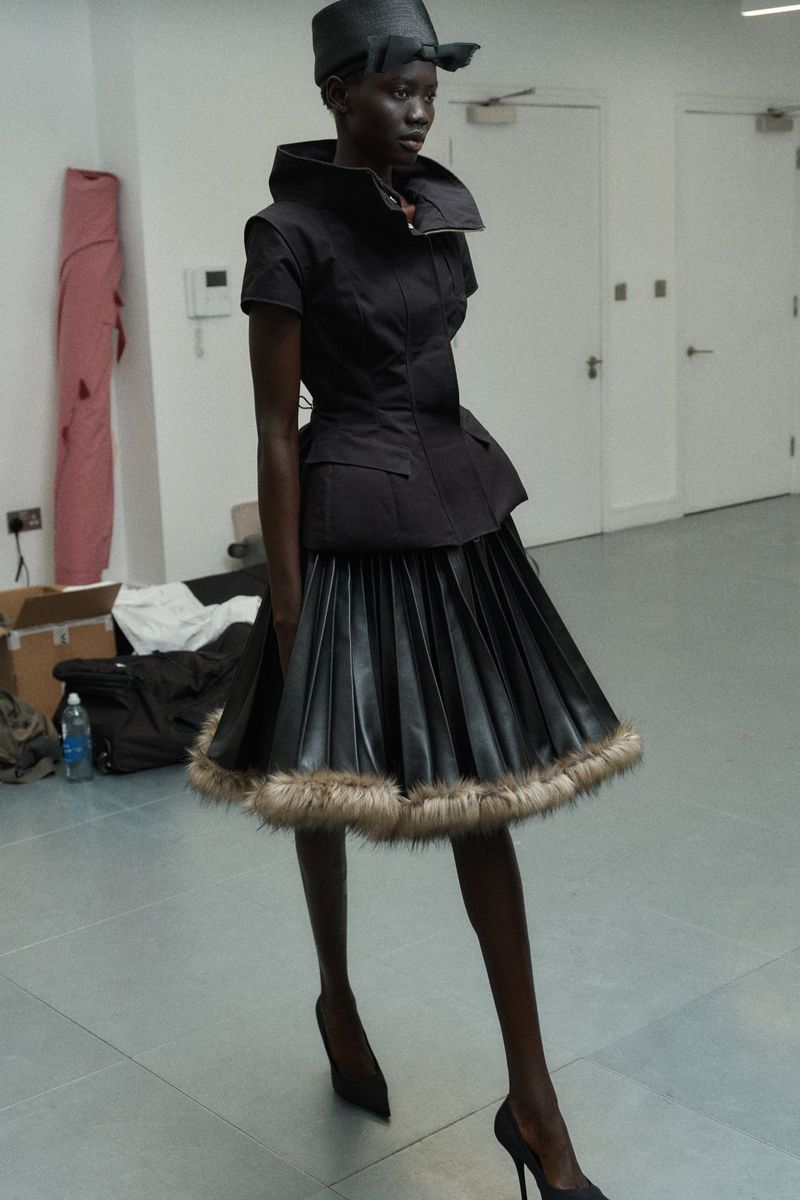 A slender Black model showcases a black peplum top and black pleated skirt with fur trim, paired with black heels in a studio setting.