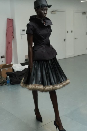 A slender Black model showcases a black peplum top and black pleated skirt with fur trim, paired with black heels in a studio setting.