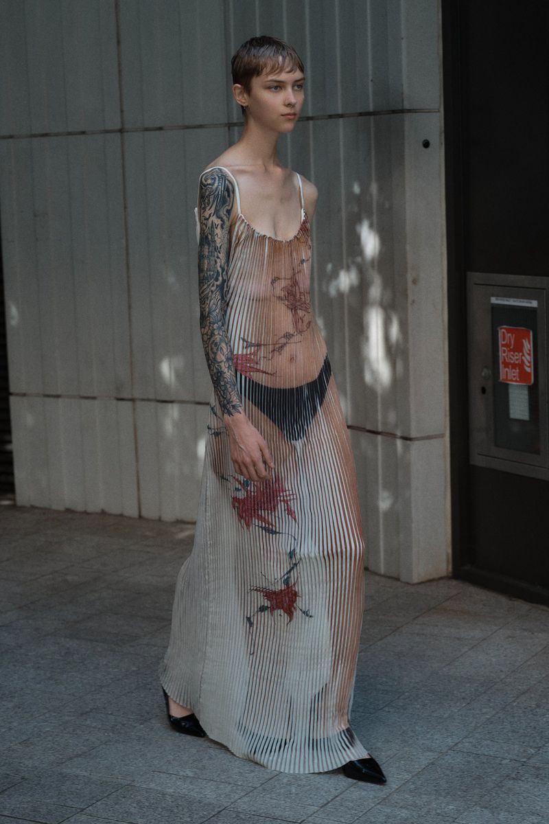 Fashionable adult female walking in a sheer dress with floral patterns on a city street with gray concrete walls in daylight.