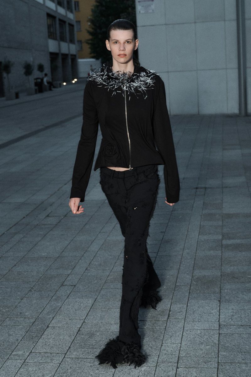 An adult female walks on a concrete surface wearing a black jacket with decorative collar and black trousers with fringes.