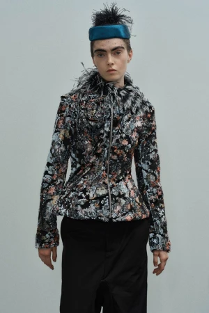 Fashion portrait of a woman wearing a floral jacket with fur collar, black pants, and teal hat against a neutral background in studio lighting.