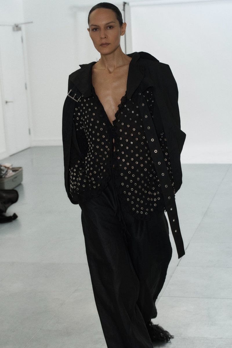 A fashion photo showcasing an adult woman in a black outfit with silver grommets, posing in a bright white studio setting.