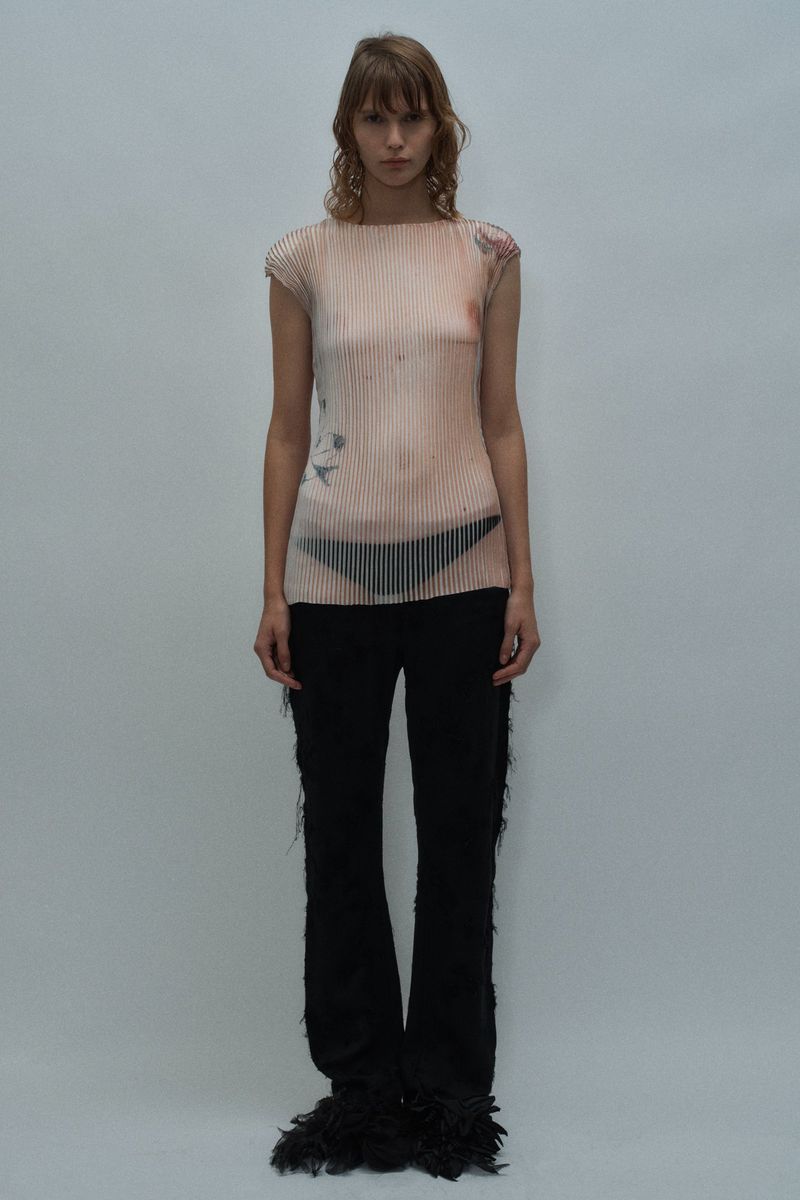 Model in a nude trompe l'oeil printed top and black fringed flare trousers standing against a pale grey studio background.