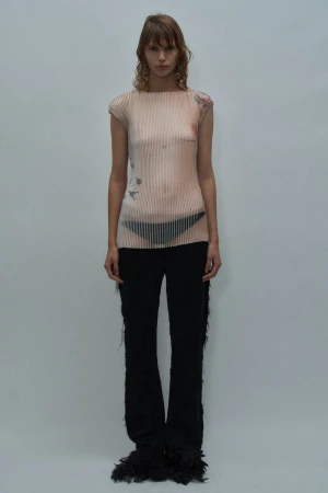 Model in a nude trompe l'oeil printed top and black fringed flare trousers standing against a pale grey studio background.