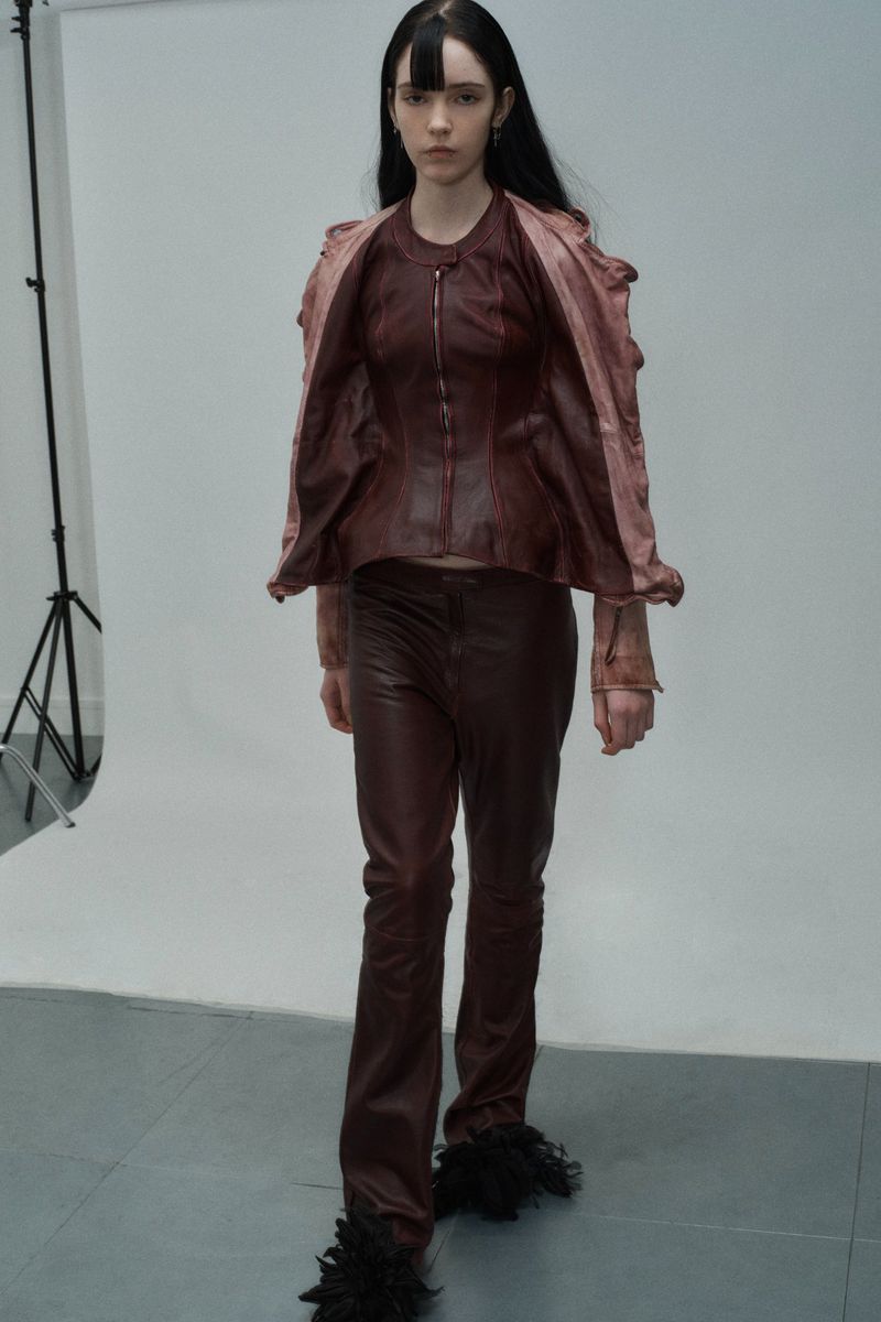 Adult fashion model in a burgundy leather jacket and pants, walking in a studio setting with natural lighting and a white background.